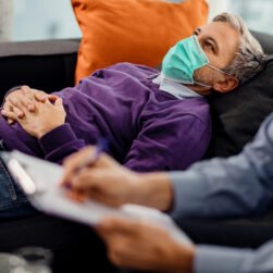 Thoughtful man with face mask lying down on psychiatrist's couch during a therapy session with mental health professional.